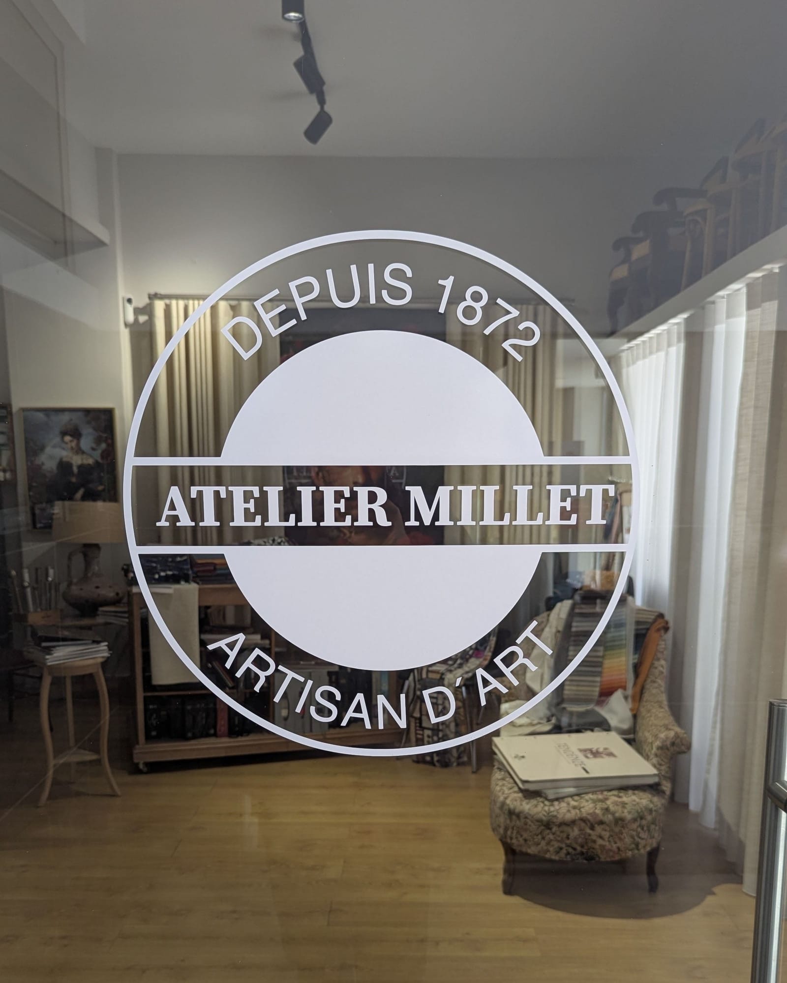 Porta de vidro do Atelier Millet com o logótipo; atrás, o showroom com um cadeirão, catálogos de tecidos e um quadro